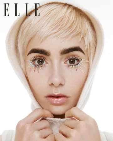 Close-up portrait of a woman with short blonde hair and hooded white top wearing colourful spiked lower eyelashes against white background.