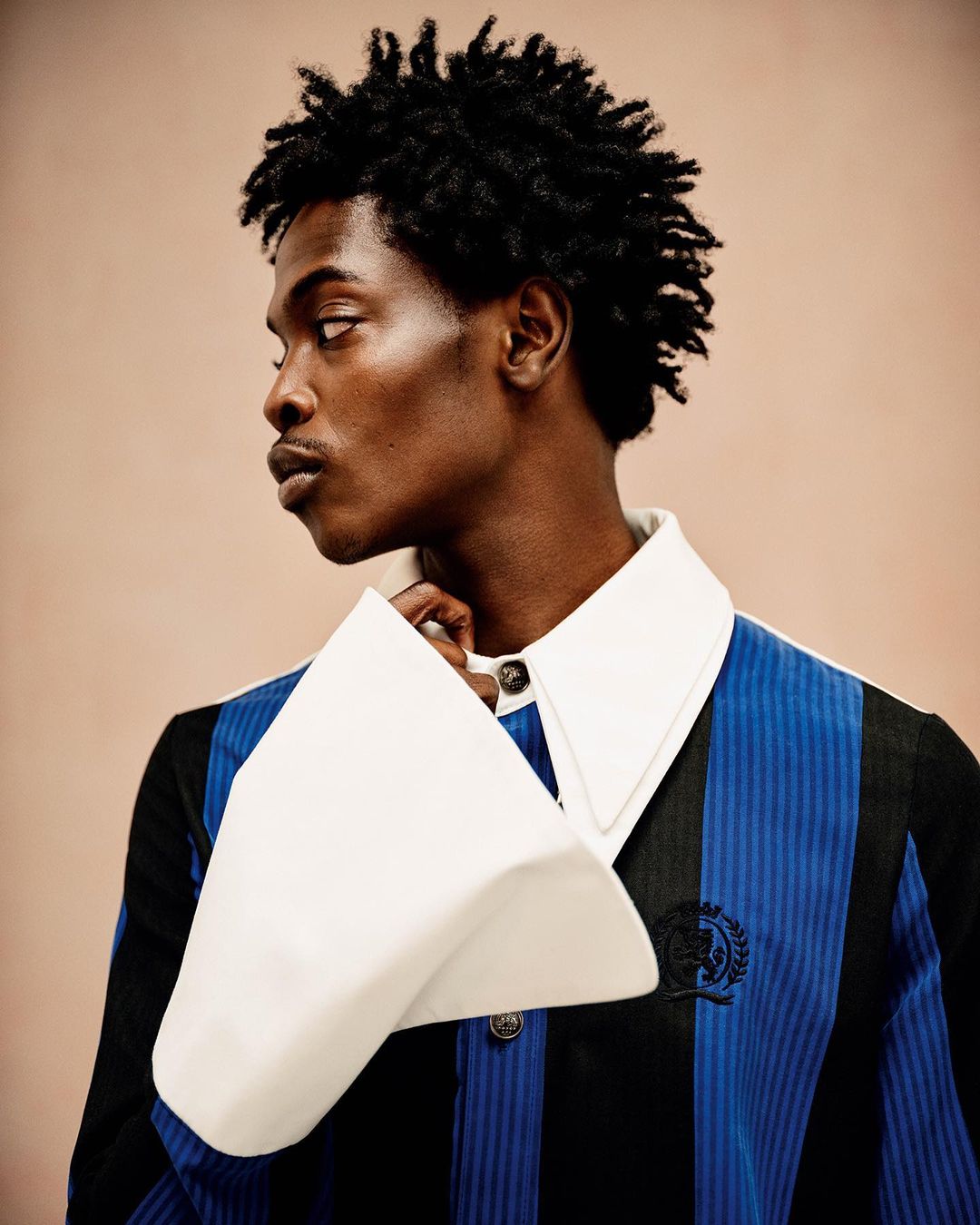 Portrait of a Black man with dark curly hair in a blue and black striped shirt with large white cuffs and collar, looking to the side.