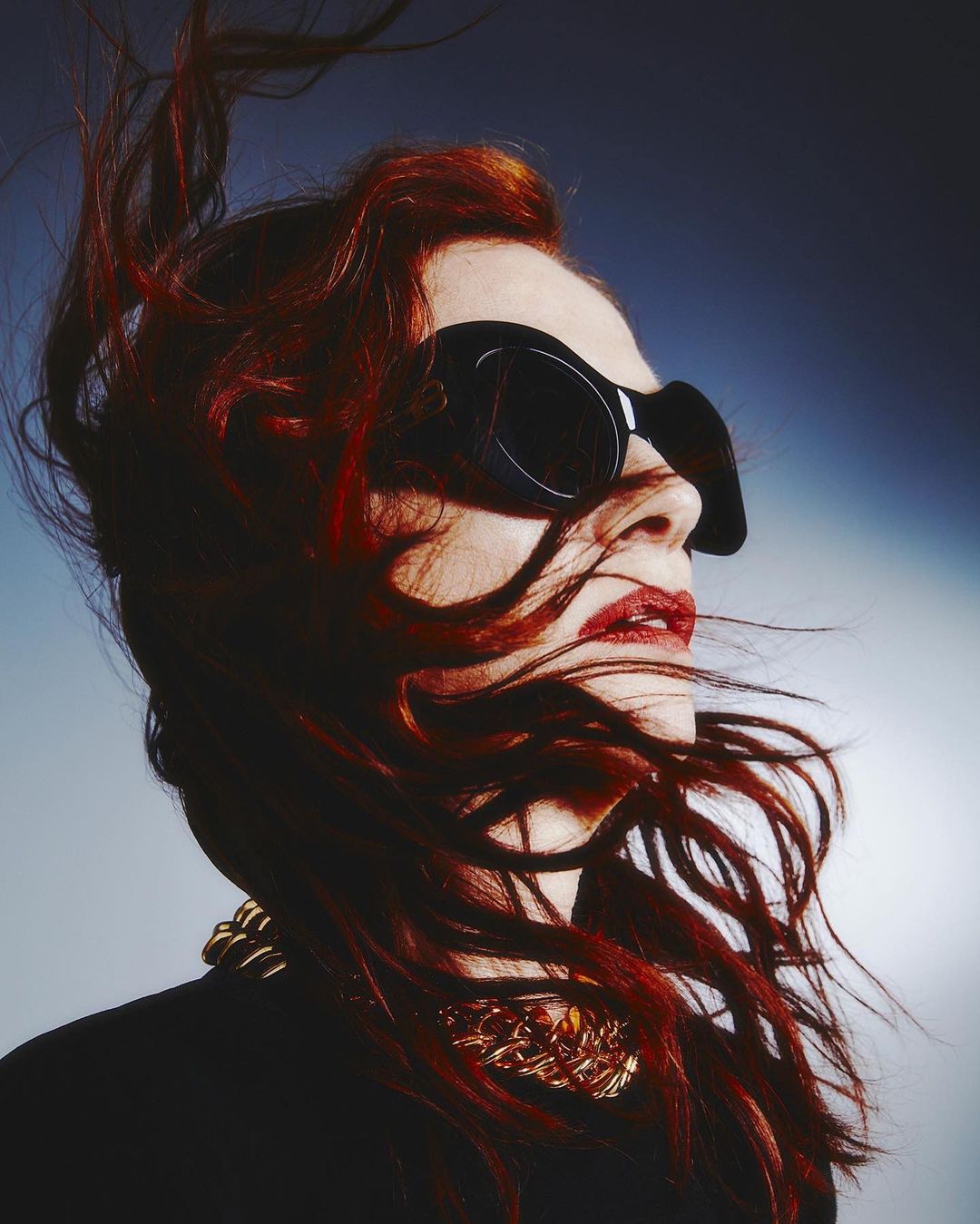 Portrait of a woman with red hair wearing oversized black sunglasses and a gold necklace against a gradient background.