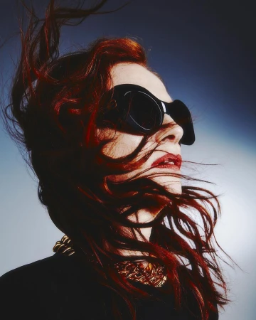 Portrait of a woman with red hair wearing oversized black sunglasses and a gold necklace against a gradient background.
