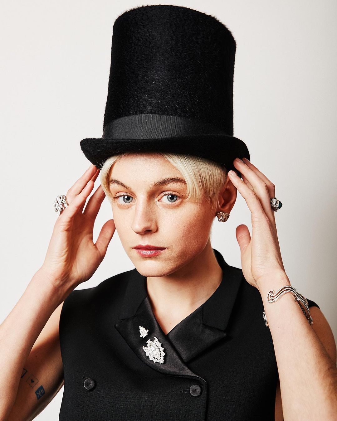 Portrait of a fair-skinned person wearing a black top hat and vest with large diamond jewelry against a neutral background.