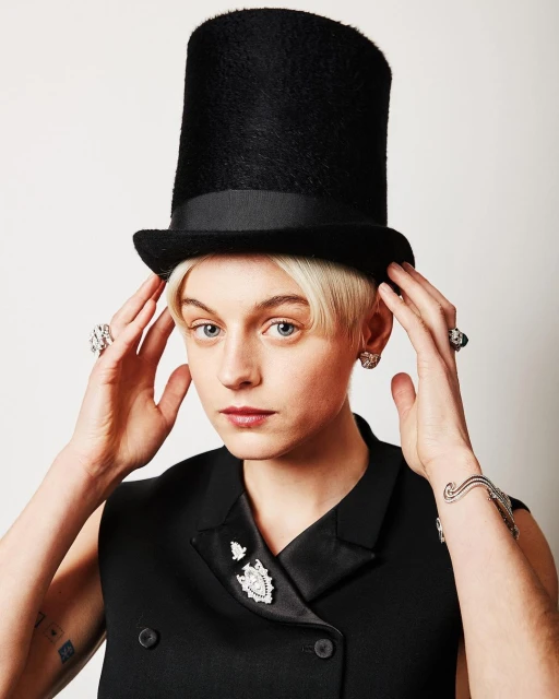 Portrait of a fair-skinned person wearing a black top hat and vest with large diamond jewelry against a neutral background.