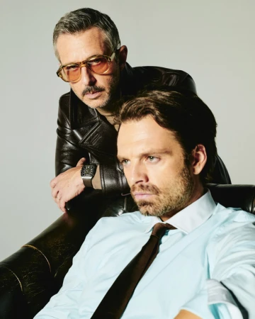 Two men posing together against a plain white background, one seated wearing a light blue dress shirt and brown tie, the other standing behind in a dark leather jacket and tinted glasses.