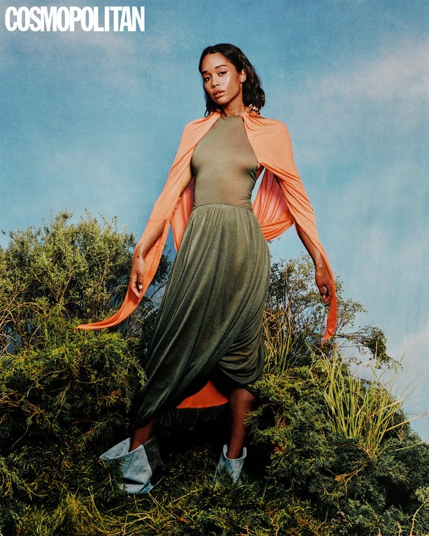 Fashion editorial shot of an adult woman in a green dress and peach shawl posing on a grassy hill under a blue sky. Magazine cover.