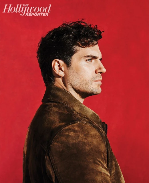 Profile portrait of actor Henry Cavill against a red background, wearing a brown suede jacket, and featuring text from The Hollywood Reporter.
