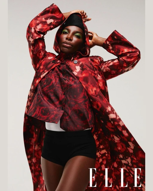 A fashionable portrait of a black woman in a hooded jacket, paired with a top and black shorts, set against a plain backdrop. ELLE logo.