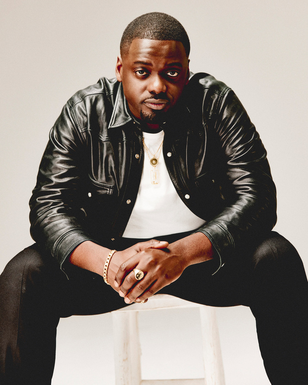 Portrait of actor Daniel Kaluuya in a black leather jacket, seated on a white stool against a neutral background, looking at camera.