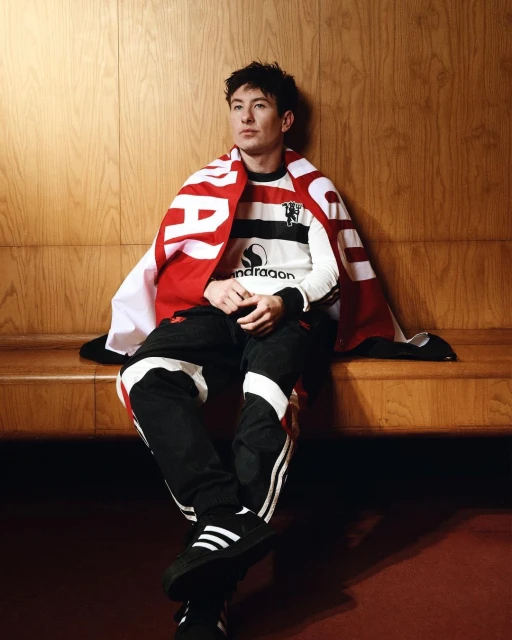 Adult male in Manchester United jersey sitting indoors, wearing Adidas track pants and sneakers, wrapped in branded red and white scarf.