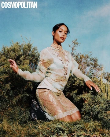 Woman in sheer embroidered high-neck top and skirt kneeling amongst green bushes beneath a blue sky