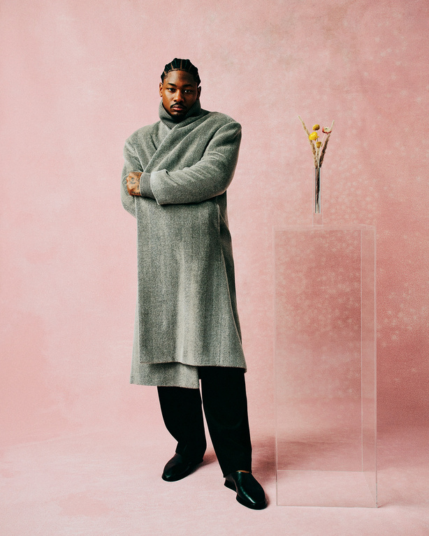 Full-length portrait of a Black man with braided hair wearing a gray wrap coat and black pants, standing in front of a pink background.