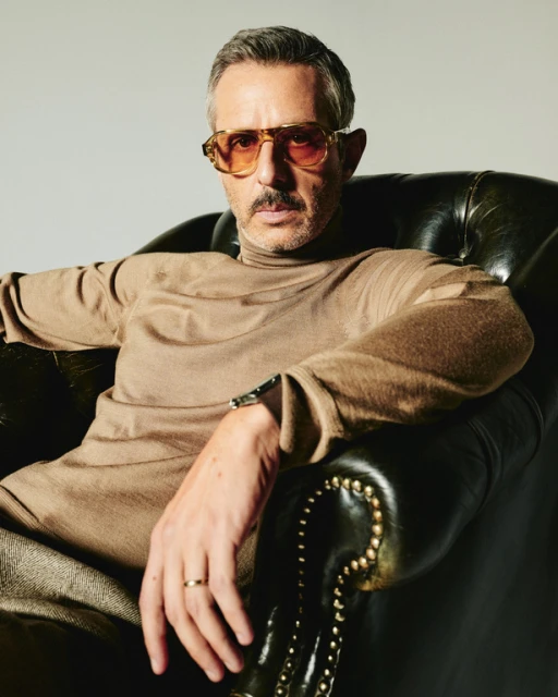 An adult male with a mustache and glasses reclines in a leather armchair wearing a tan turtleneck sweater, wrist watch, and wedding ring.