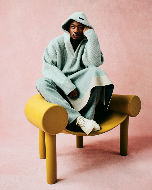 An adult male wearing a hooded robe and sneakers sits on a yellow chair in a portrait shot against a solid pink background.