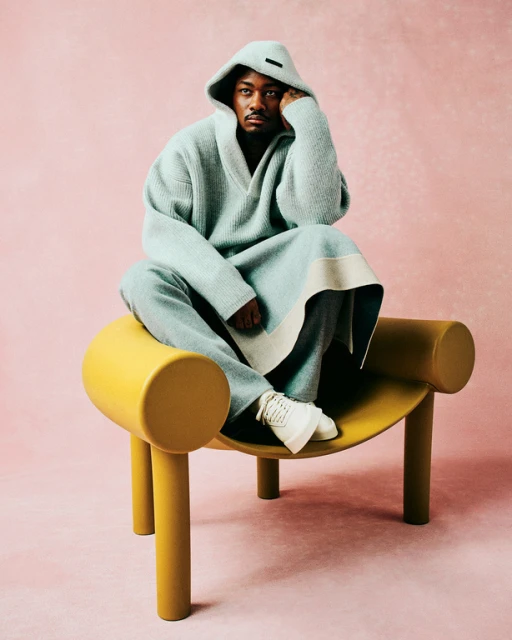 An adult male wearing a hooded robe and sneakers sits on a yellow chair in a portrait shot against a solid pink background.