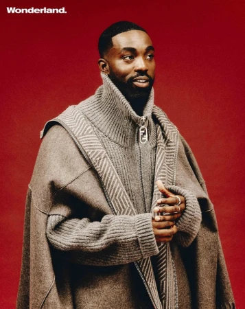 Man wearing a grey ribbed knit sweater and brown cape against a solid red background