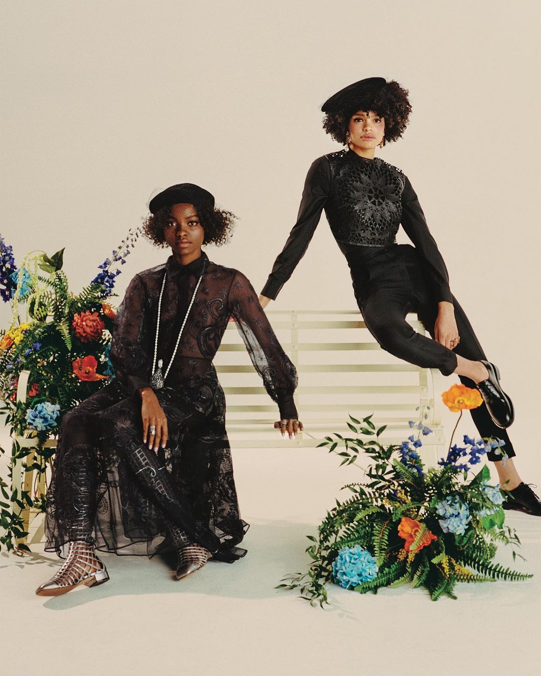 Two Black women in fashionable dark-tone clothing pose on a white bench with floral accents in a studio setting with soft, diffused lighting.