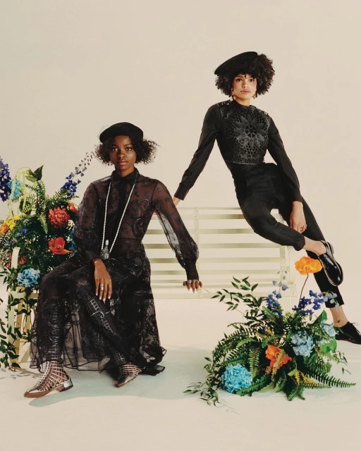 Two Black women in fashionable dark-tone clothing pose on a white bench with floral accents in a studio setting with soft, diffused lighting.