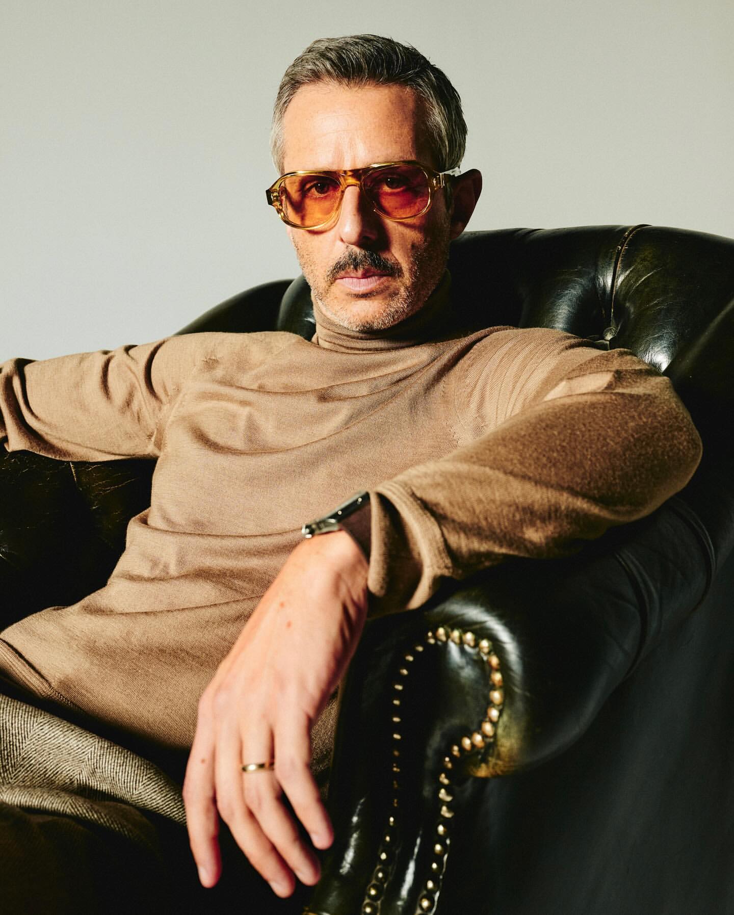 Portrait of a man with graying hair and a mustache wearing orange sunglasses, a tan turtleneck sweater, and sitting in a dark leather chair.