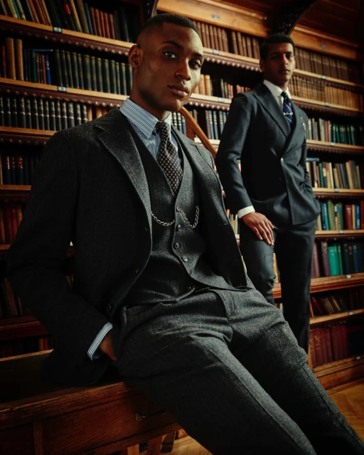 Two dark-skinned adult men in suits pose inside a library with wood bookshelves and a wooden desk. One man sits on the desk.