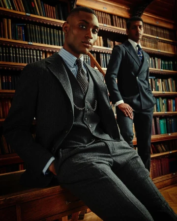 Two dark-skinned adult men in suits pose inside a library with wood bookshelves and a wooden desk. One man sits on the desk.