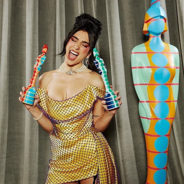 Dua Lipa poses with a statue-shaped award in a yellow dress with black netting, smiling with her mouth open, at an event.