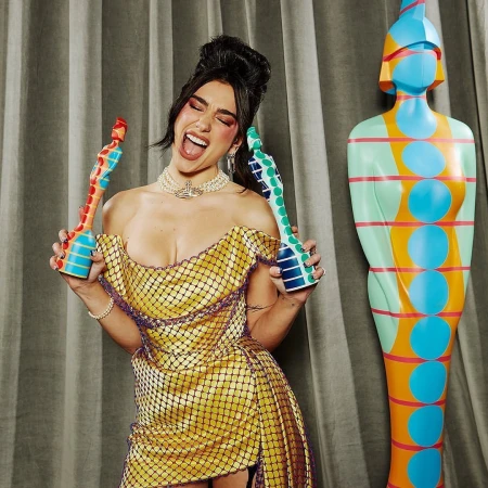 Dua Lipa poses with a statue-shaped award in a yellow dress with black netting, smiling with her mouth open, at an event.