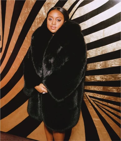 Woman wearing a large black faux fur coat posing in front of a striking black and gold striped starburst background.