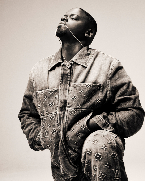 Portrait of African American male in denim jacket and pants featuring Louis Vuitton logos against neutral backdrop in a studio.