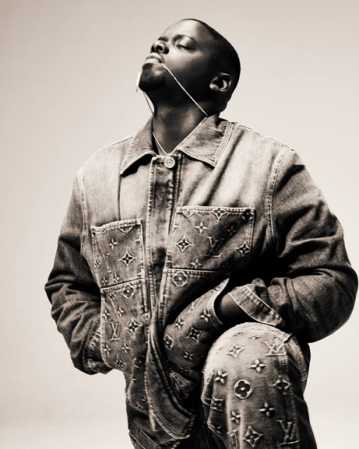 Portrait of African American male in denim jacket and pants featuring Louis Vuitton logos against neutral backdrop in a studio.