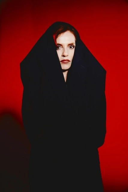 Striking portrait of a woman with red hair wrapped in black fabric against a bold red backdrop, creating a dramatic and mysterious atmosphere.