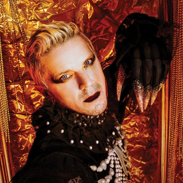 Adult male with blond hair and dramatic eye makeup poses in front of a textured gold background while wearing black clothing and pearls.