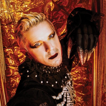 Adult male with blond hair and dramatic eye makeup poses in front of a textured gold background while wearing black clothing and pearls.
