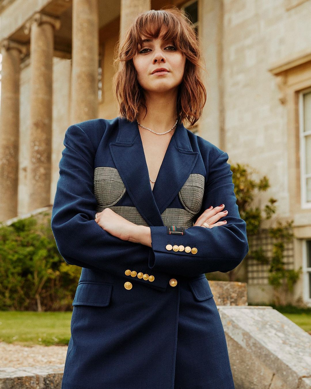 A woman poses in a blue blazer with gold buttons outdoors, set against an old building with columns, capturing a stylish, confident portrait.