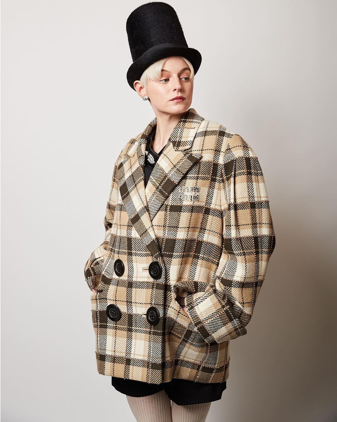 Woman in plaid jacket and top hat poses against a white backdrop, showcasing fashion and studio photography with natural light.