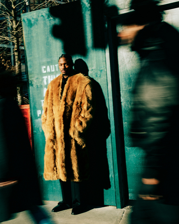 Adult male in a fur coat poses outdoors near a weathered green wall, with blurred figures passing by, capturing an urban fashion moment.