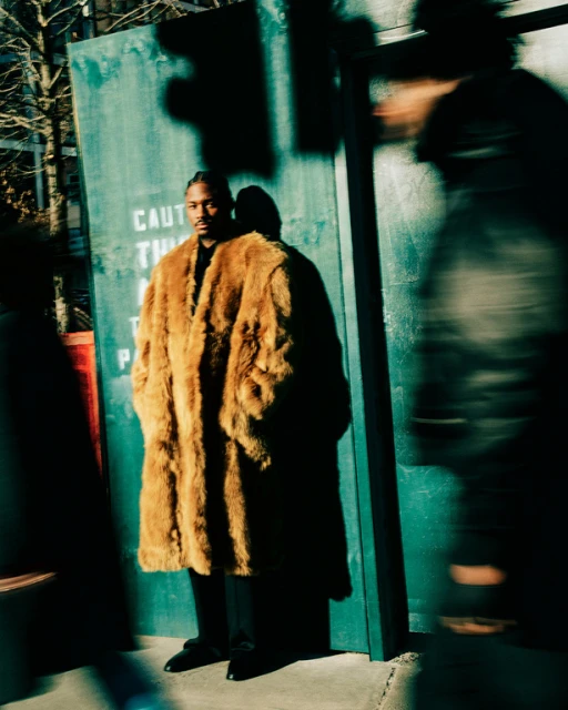Adult male in a fur coat poses outdoors near a weathered green wall, with blurred figures passing by, capturing an urban fashion moment.