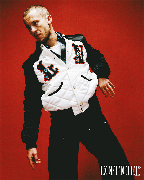 Man in black pants and a white jacket with black sleeves poses in front of a red background. 'L'OFFICIEL' logo is in the lower right corner.