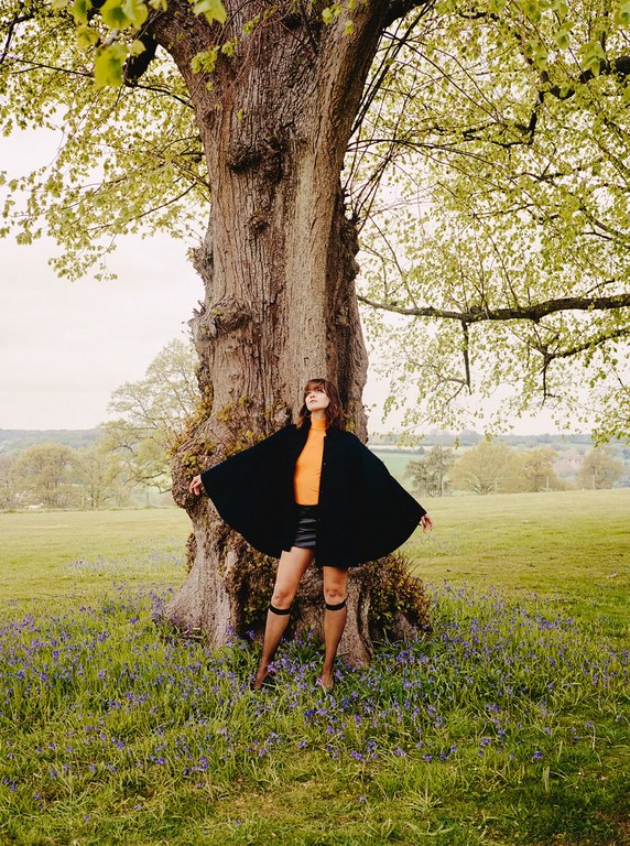 Woman in cape poses near tree in wildflower meadow. Fashion shot in natural light, with rural, outdoor setting, and neutral sky.