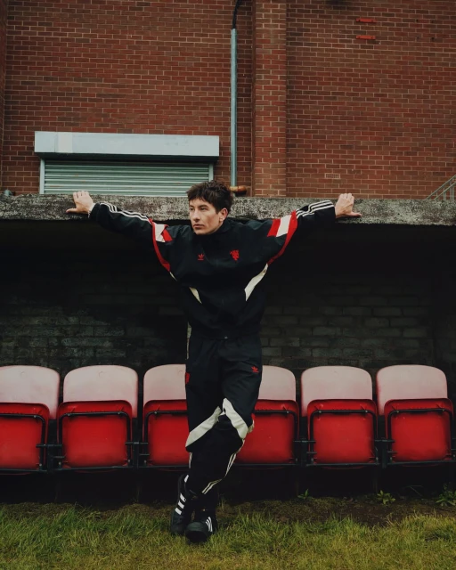 Adult male poses in black and red Adidas tracksuit against a red brick wall and stadium seating, capturing a casual, sporty aesthetic.