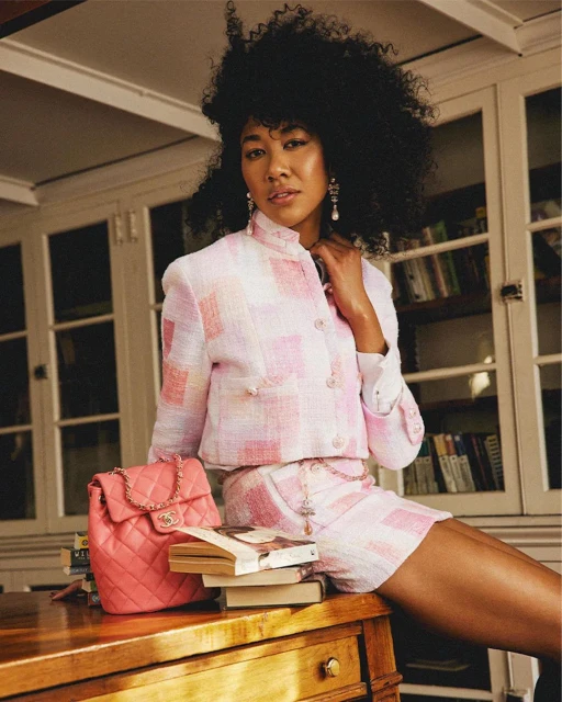 Woman in pink plaid jacket and shorts sitting on a wooden desk next to books and a pink quilted backpack in an indoor setting.
