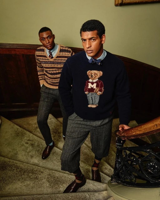 Two adult men on a staircase wearing sweaters, trousers, and dress shoes in an indoor setting with neutral-toned walls and dark wood trim.