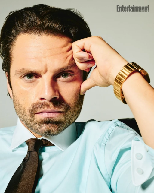 Headshot of adult male celebrity, Sebastian Stan, wearing a light blue button-down shirt and brown tie, with a gold wristwatch.