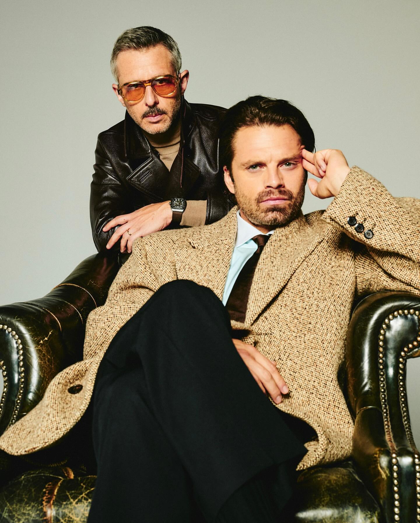 Two adult males pose indoors: one seated in a leather chair in a beige coat and black pants, the other standing in a leather jacket.