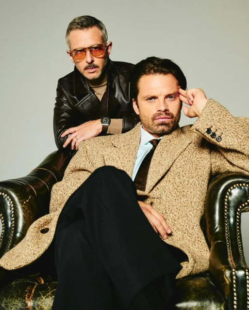 Two adult males pose indoors: one seated in a leather chair in a beige coat and black pants, the other standing in a leather jacket.