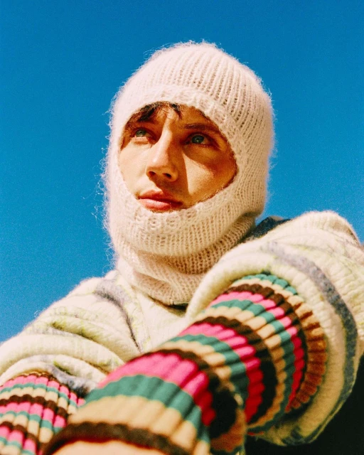 Eye-level portrait of an adult male wearing a knitted balaclava and striped knit sweater against a bright blue sky background.