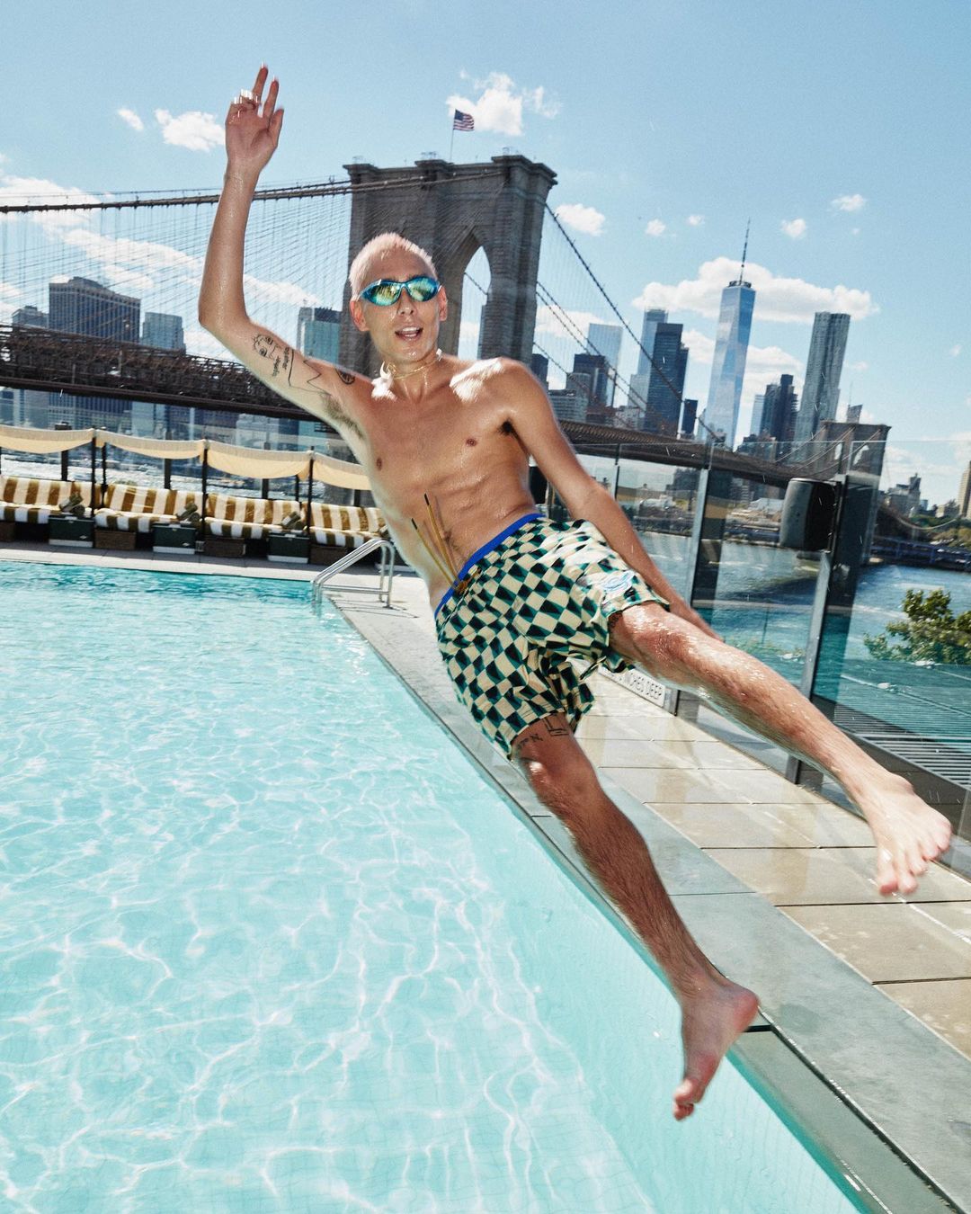 An adult male with pink hair wearing swimming goggles and patterned shorts jumping into a swimming pool with a city skyline in the background.