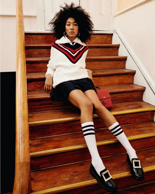 Attractive woman with afro sits on wooden stairs, wearing a white sweater, blue skirt, striped socks, and black loafers with a red purse.