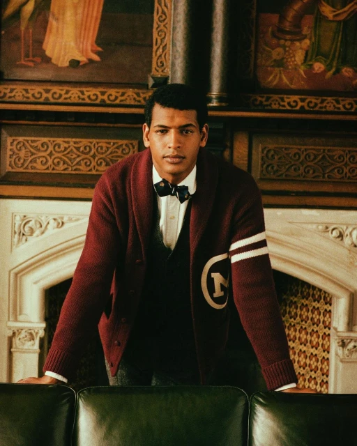 Young man in cardigan, bow tie, and vest posing indoors with ornate woodwork and decorative painting in the background.