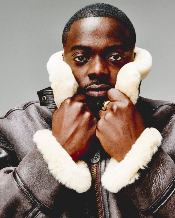 Close-up portrait of a Black man with short hair wearing a brown shearling coat, hands up to the collar against a grey backdrop.