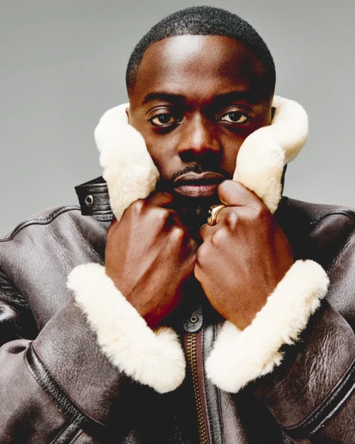 Close-up portrait of a Black man with short hair wearing a brown shearling coat, hands up to the collar against a grey backdrop.