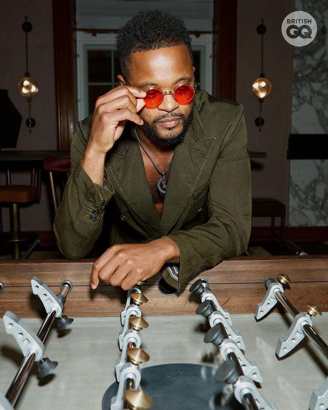 Portrait of an adult Black male wearing an olive-green suit and red sunglasses leaning over a foosball table indoors with moody lighting.